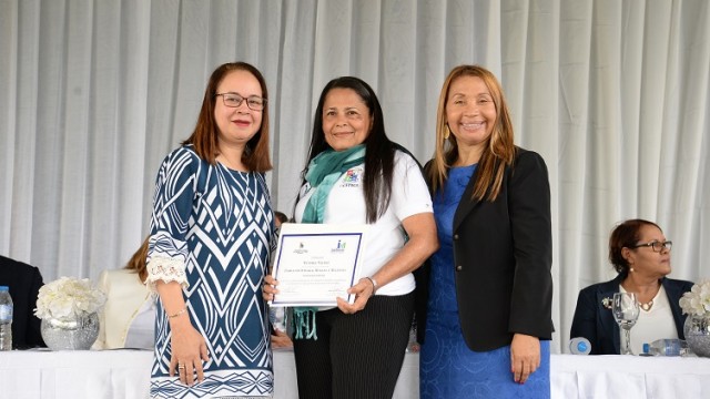 La Dra. Alina Bello y la Lic. Reyita Pinales entregan el certificado de participación a la profesora Victoria Valdez.